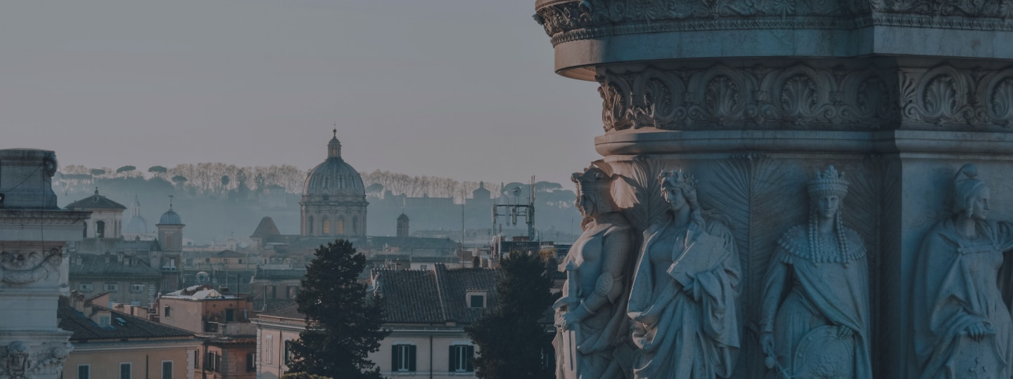 Stone statues on a historic building in the foreground with rooftops and a domed church in the background in a European city.