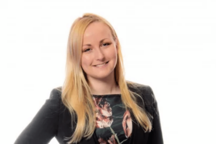 A woman with long blonde hair, wearing a black blazer and a floral patterned top, smiles at the camera against a plain white background.