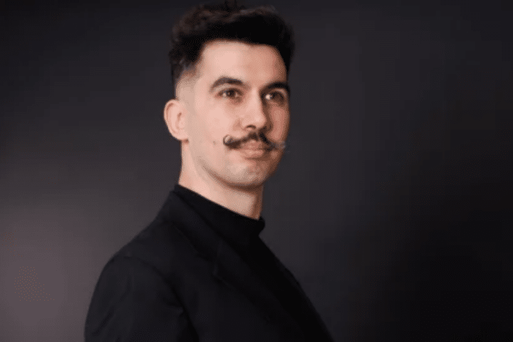 Man with dark hair, styled moustache, and a black jacket poses against a dark, plain background, looking slightly to the side.