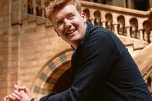 A person with short red hair wearing a dark shirt is smiling and sitting in front of a stone staircase with arched railings.