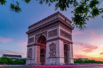 The Arc de Triomphe in Paris is shown at sunset with a colourful sky and tree branches framing the top of the image.