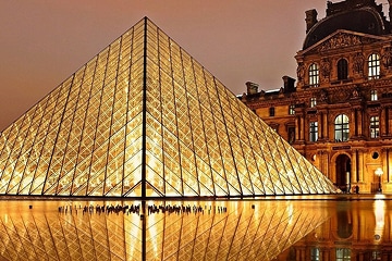 The illuminated glass pyramid entrance of the Louvre Museum in Paris is reflected in a pool of water at night, with part of the historic building visible on the right.
