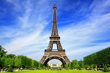 The Eiffel Tower stands against a blue sky with green trees and grass on either side, viewed from the park below.