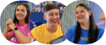Three young people smile and talk while sitting indoors, each wearing brightly coloured shirts—pink, yellow, and blue—against a blurred background.