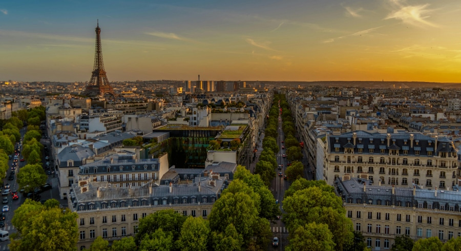 Aerial view of Paris at sunset, showing the Eiffel Tower on the left, tree-lined streets, and classic Parisian buildings.