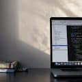 A laptop displaying code sits on a desk next to a stack of books, with soft light casting shadows on the wall behind.
