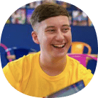 A young person with short brown hair, wearing a yellow shirt, smiles whilst seated indoors with colourful chairs and photos in the background.