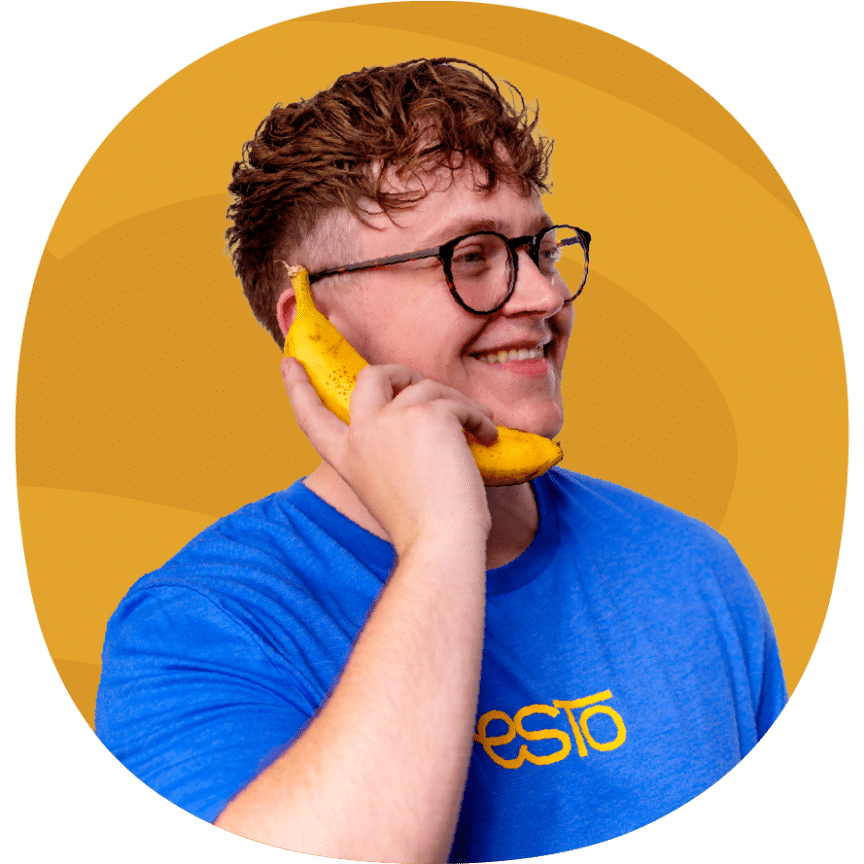 A person wearing glasses and a blue shirt holds a banana to their ear as if using it as a phone, smiling in front of an orange background.