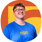 Person with short curly hair and glasses, wearing a blue shirt with “esto” text, standing in front of a yellow and orange background.