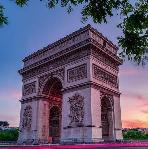 The Arc de Triomphe in Paris, photographed at sunset with a pink and purple sky, framed by tree branches at the top.
