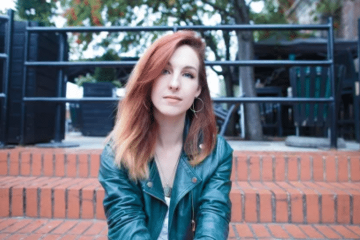 Woman with red hair wearing a green leather jacket sits on outdoor brick steps with trees and a railing in the background.