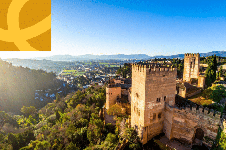 A wide view of the Alhambra fortress in Granada, Spain, with surrounding greenery and hills under a clear blue sky; a yellow abstract logo overlays the top left corner.