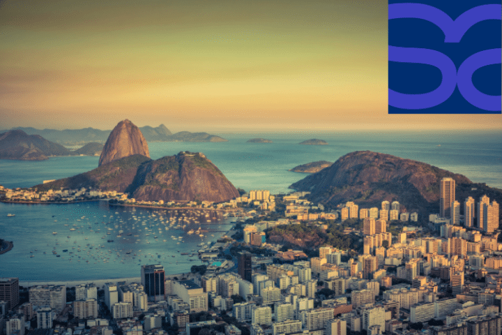 A cityscape of Rio de Janeiro at sunset, featuring Sugarloaf Mountain and Guanabara Bay, with a blue abstract logo in the top right-hand corner.
