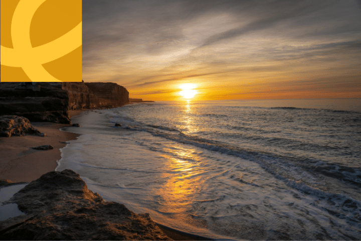 A sunset over the ocean with gentle waves on a sandy beach and rocky shoreline; a yellow geometric logo is overlaid in the top left corner.