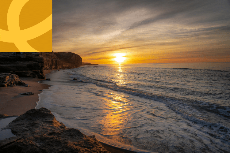 A sunset over the ocean with gentle waves on a sandy beach and rocky shoreline; a yellow geometric logo is overlaid in the top left corner.