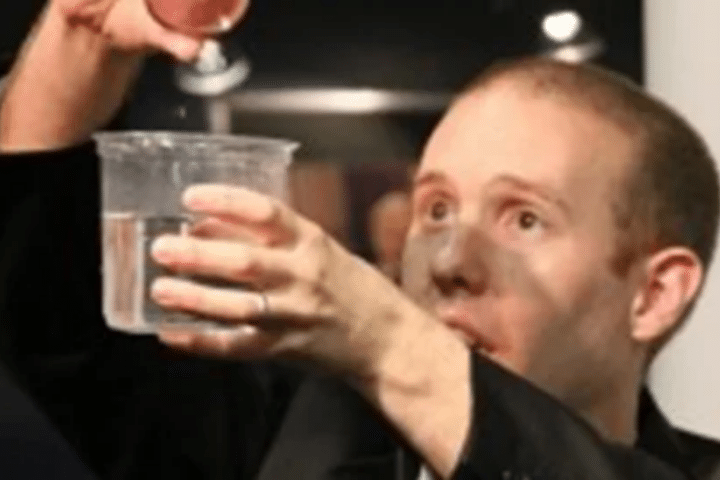 A person holds a clear plastic cup of water in one hand and appears to sprinkle or add something into it with the other hand.