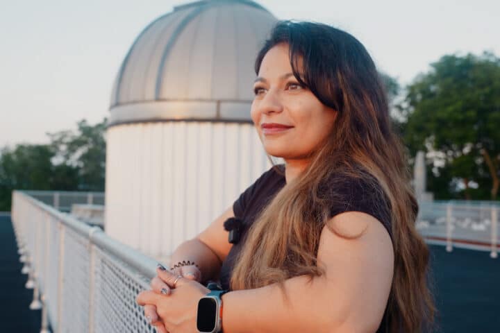 A woman with long brown hair stands by a fence, looking into the distance, with a domed observatory building in the background.