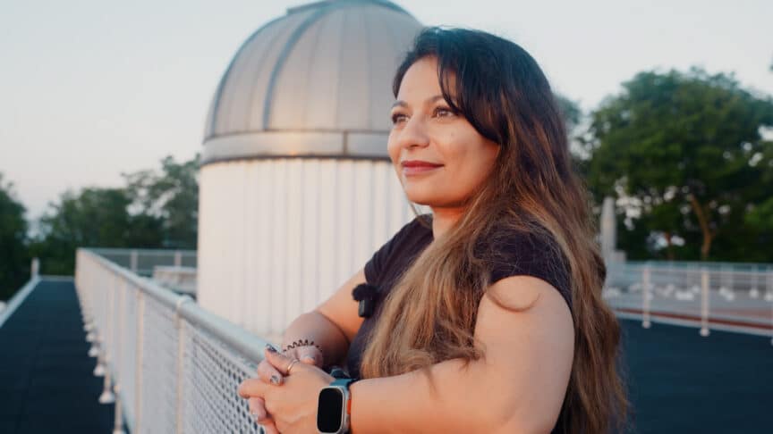 A woman with long brown hair stands by a fence, looking into the distance, with a domed observatory building in the background.