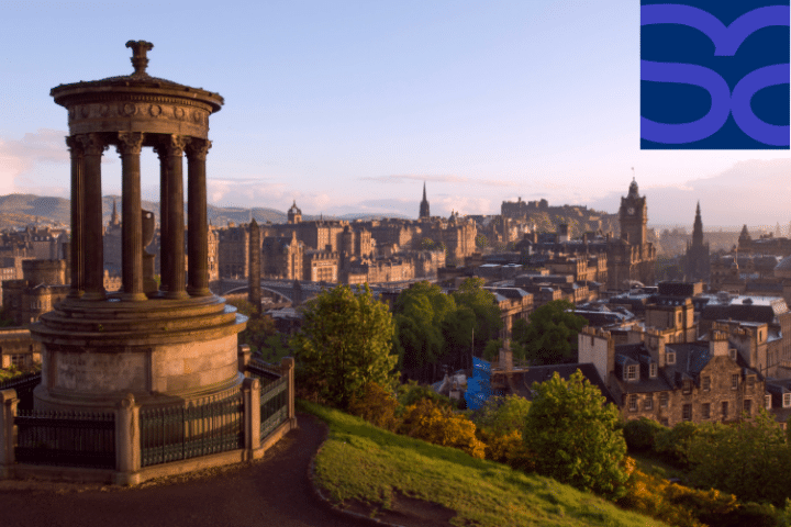 View of Edinburgh cityscape from Calton Hill at sunset, featuring the Dugald Stewart Monument in the foreground and historic buildings in the background. A purple and blue logo is in the top right-hand corner.