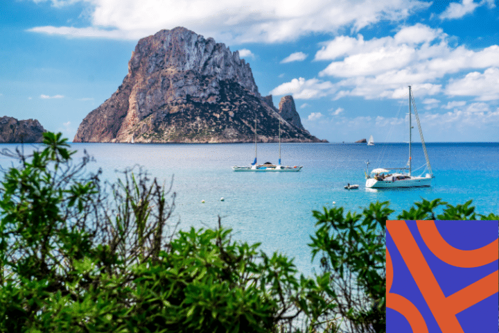 A rocky island rises from blue sea under a partly cloudy sky, with sailboats anchored near shore and green shrubs in the foreground. A colourful abstract pattern appears in the corner.