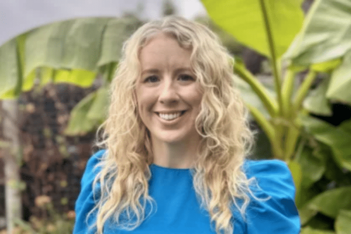 A woman with long curly blonde hair, wearing a bright blue top, stands outdoors in front of large green banana leaves.