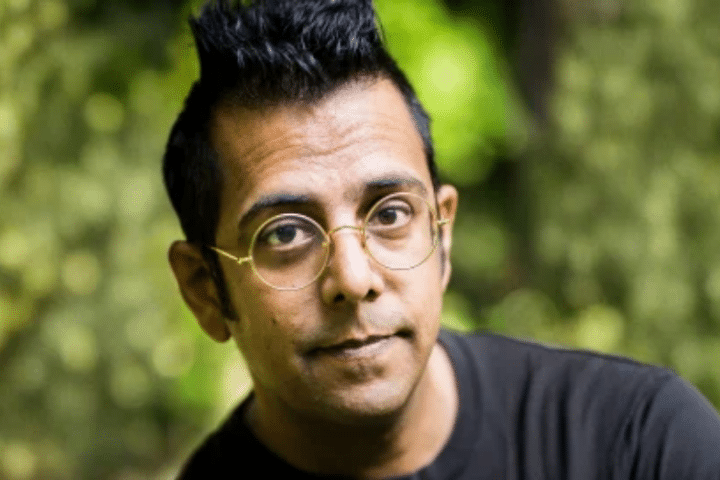 A person with short black hair, wearing round glasses and a dark shirt, is looking at the camera with a neutral expression, outdoors with greenery in the background.