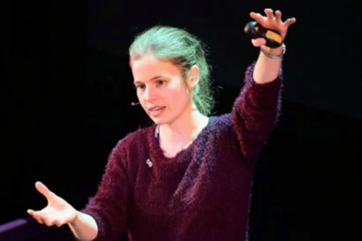 A person with long hair tied back gestures with both arms while speaking onstage, wearing a dark jumper and holding a small object in one hand.