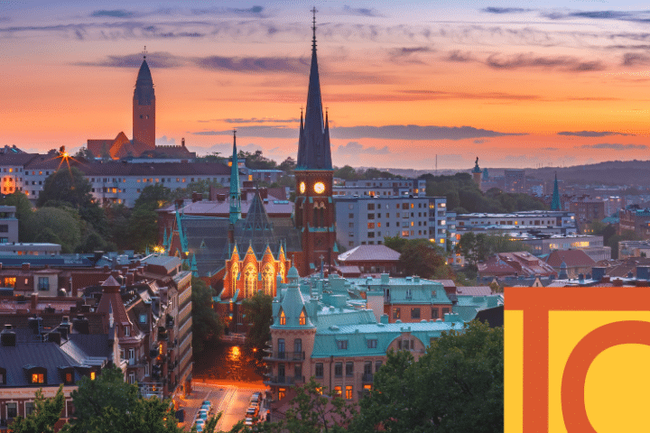 A cityscape at sunset featuring historic buildings, a tall church spire with a clock, and colourful rooftops under a partly cloudy sky.