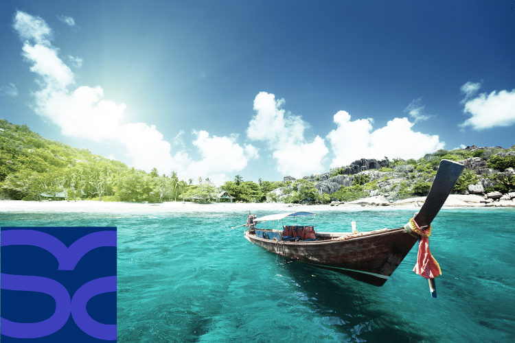 A wooden boat floats on clear turquoise water near a tropical island with lush greenery under a sunny sky; a blue logo appears in the lower left corner.