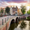 A stone bridge crosses a canal lined with trees and traditional Dutch buildings under a colourful sky at sunset in Amsterdam.