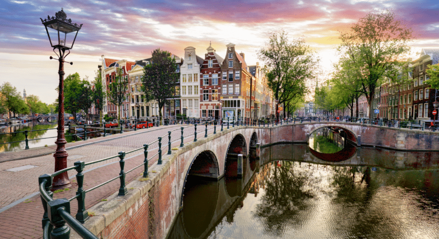 Arched brick bridge over a canal with historic buildings, trees, and street lamps at sunset in a European city.