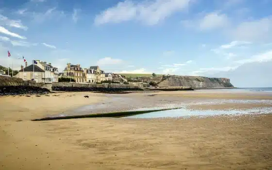 Beachfront buildings and flags near sandy shore with cliffs in the background under a partly cloudy sky.