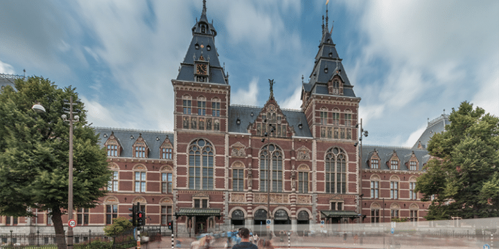 The image shows the front façade of the Rijksmuseum in Amsterdam, a large red-brick building with towers, ornate details, and a crowd of people blurred in the foreground.