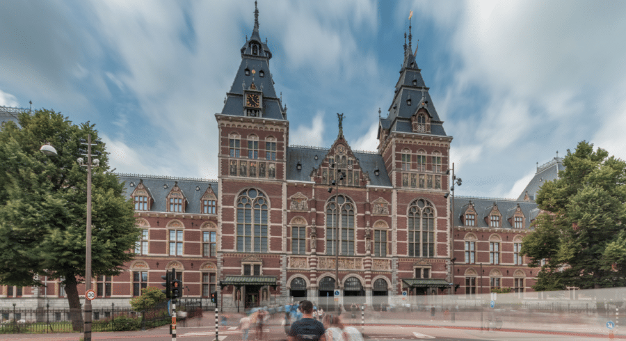 The façade of the Rijksmuseum in Amsterdam, featuring red brickwork, arched windows, and twin towers under a partly cloudy sky with blurred motion of people in front.