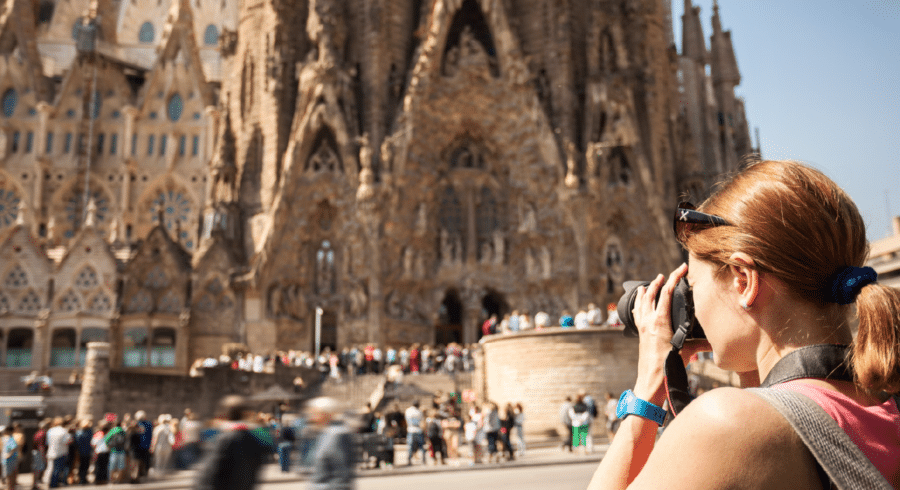 A woman in sunglasses takes a photo of a large, ornate cathedral with many people gathered outside the entrance on a sunny day.