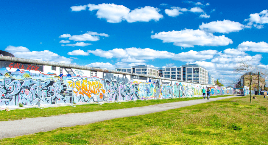 A long section of the Berlin Wall covered in colourful graffiti, with green grass, people walking, and modern buildings in the background under a blue sky with clouds.