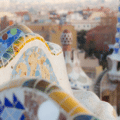 Close-up of colourful mosaic tiles on a curved bench in Park Güell, Barcelona, with city buildings and Gaudí architecture in the blurred background.