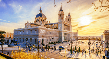 Large historic cathedral with domes and towers, set against a bright sunset sky, with many people walking in the square in front.