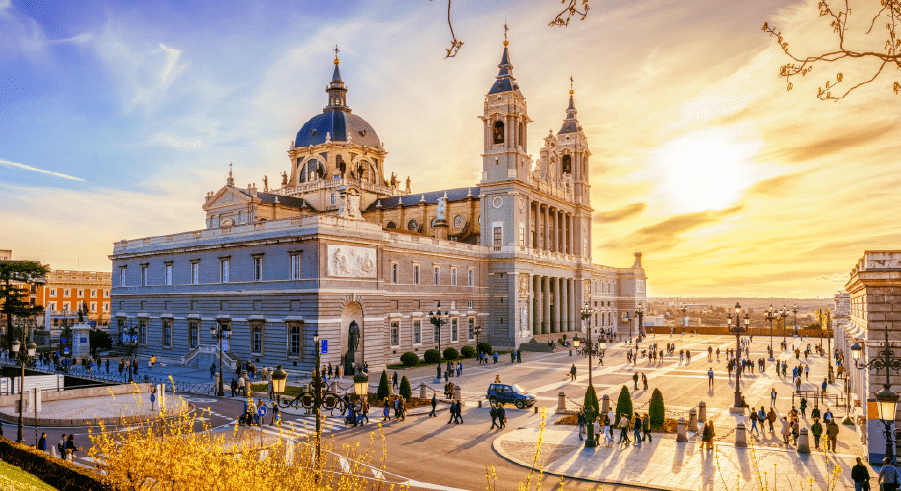 Almudena Cathedral in Madrid, Spain, with people walking in the square at sunset under a partly cloudy sky.