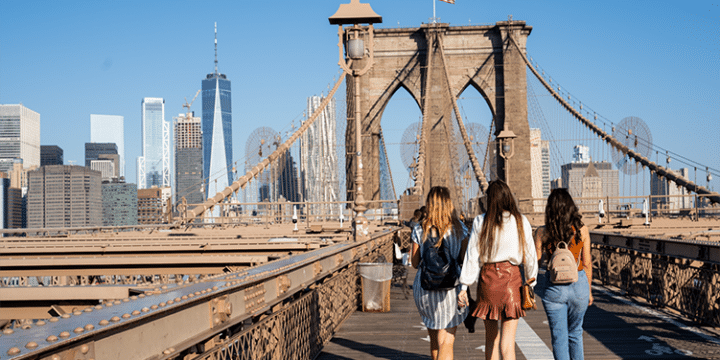 Three people walk on the Brooklyn Bridge in New York City with skyscrapers and a clear sky in the background.