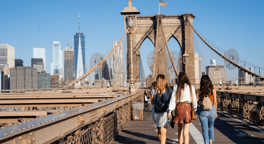 Three women walk on the Brooklyn Bridge towards Manhattan on a clear day, with city skyscrapers visible in the background.