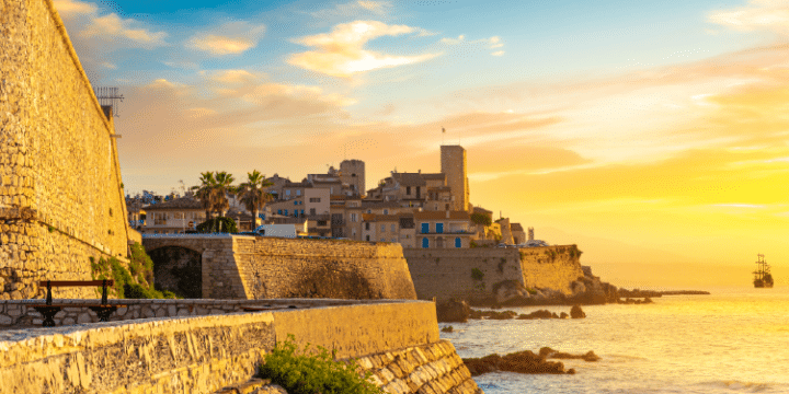 Sunset view of a coastal town with stone walls, historic buildings, palm trees, and a ship sailing on the calm sea.
