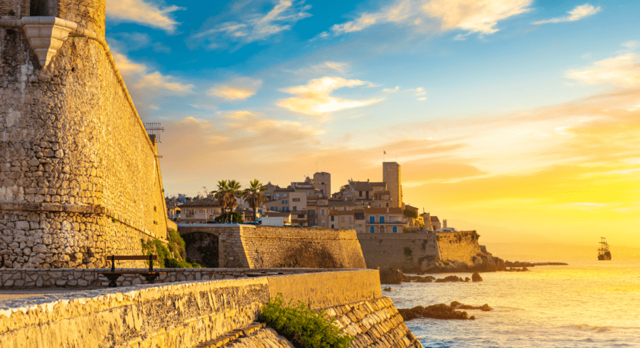 Stone walls overlook the sea at sunset, with a coastal town and a distant ship visible under a partly cloudy sky.