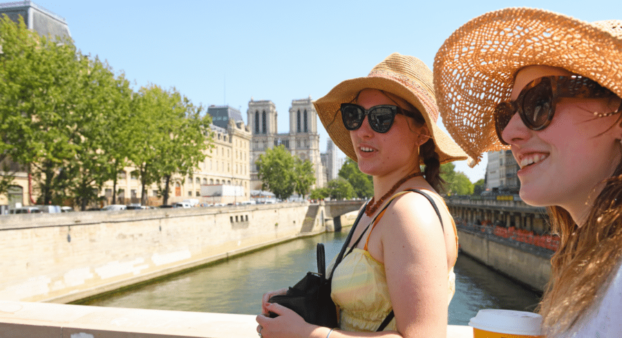 Two women wearing sunglasses and straw hats stand on a bridge over a river in a city on a sunny day, holding drinks and looking towards the water.
