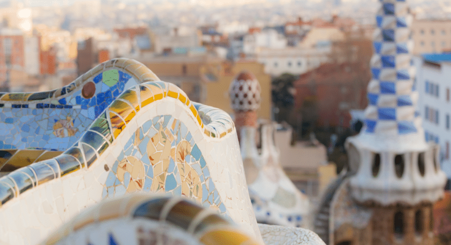 Close-up of colourful mosaic bench in Park Güell, Barcelona, with city buildings and distinctive mosaic towers blurred in the background.