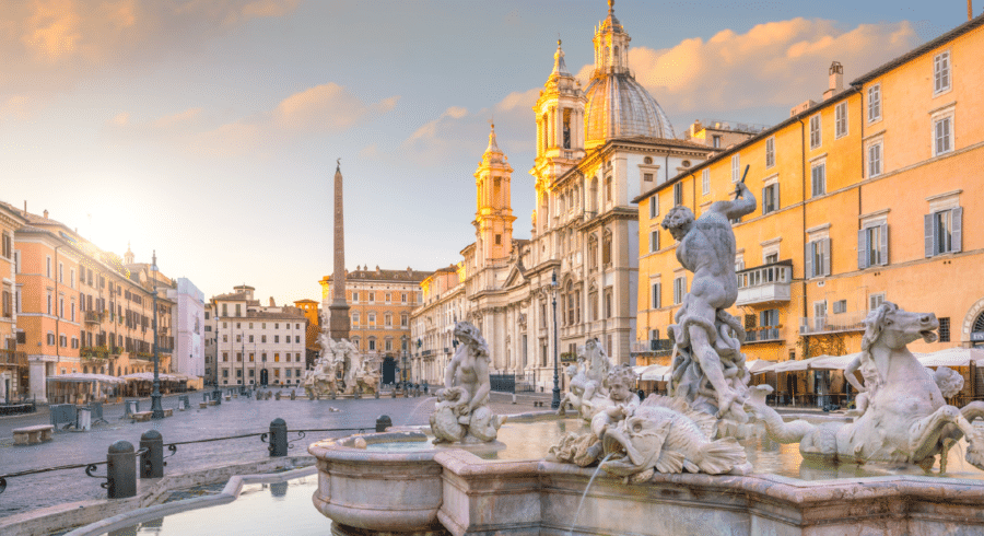 A large baroque fountain with sculptures stands in a historic city square surrounded by ornate buildings at sunrise.