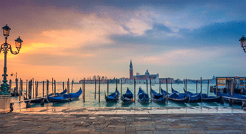 Row of gondolas moored at a pier on a calm lagoon with a distant view of an historic building and bell tower at sunrise or sunset in Venice, Italy.