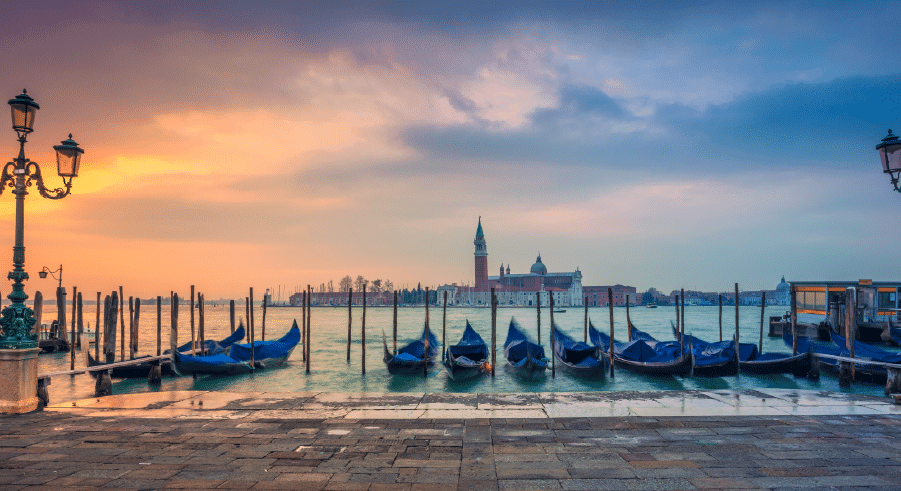 Row of covered gondolas moored at a pier on the Grand Canal in Venice at sunset, with the island of San Giorgio Maggiore and its church visible across the water.