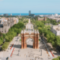 Aerial view of the Arc de Triomf in Barcelona, surrounded by trees and city buildings, with a wide promenade leading towards the sea.
