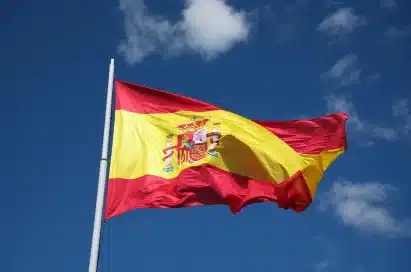 The flag of Spain flying on a flagpole against a blue sky with some clouds.